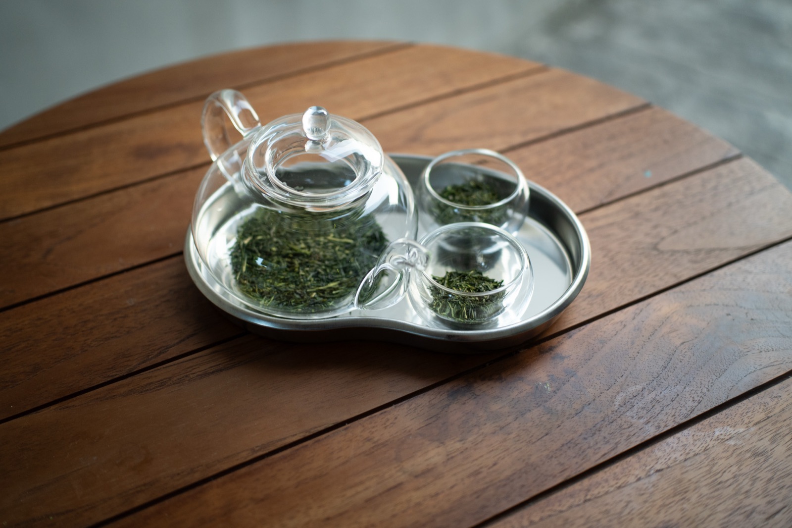 A glass teapot and two small glass cups of loose tea leaves on a steel tray, on a wooden table